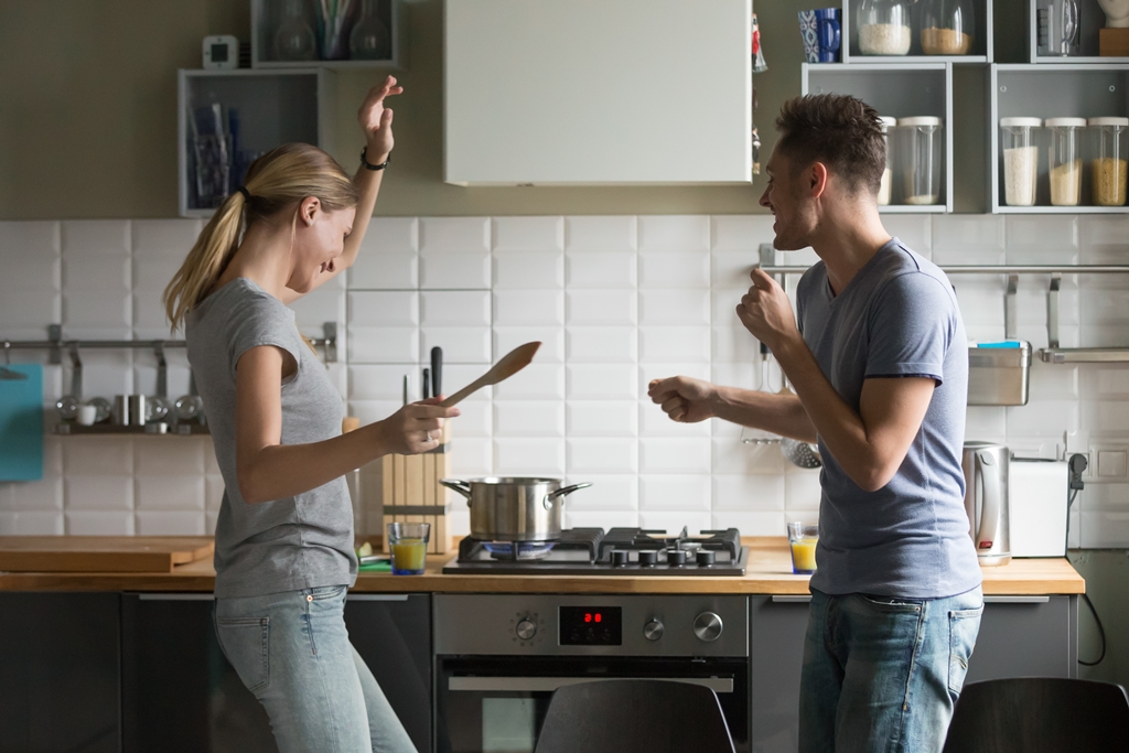 Two people playfully dancing in a kitchen with cooking utensils in hand.