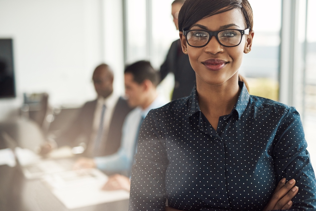 Confident businesswoman with crossed arms standing in front of colleagues in a modern office setting.