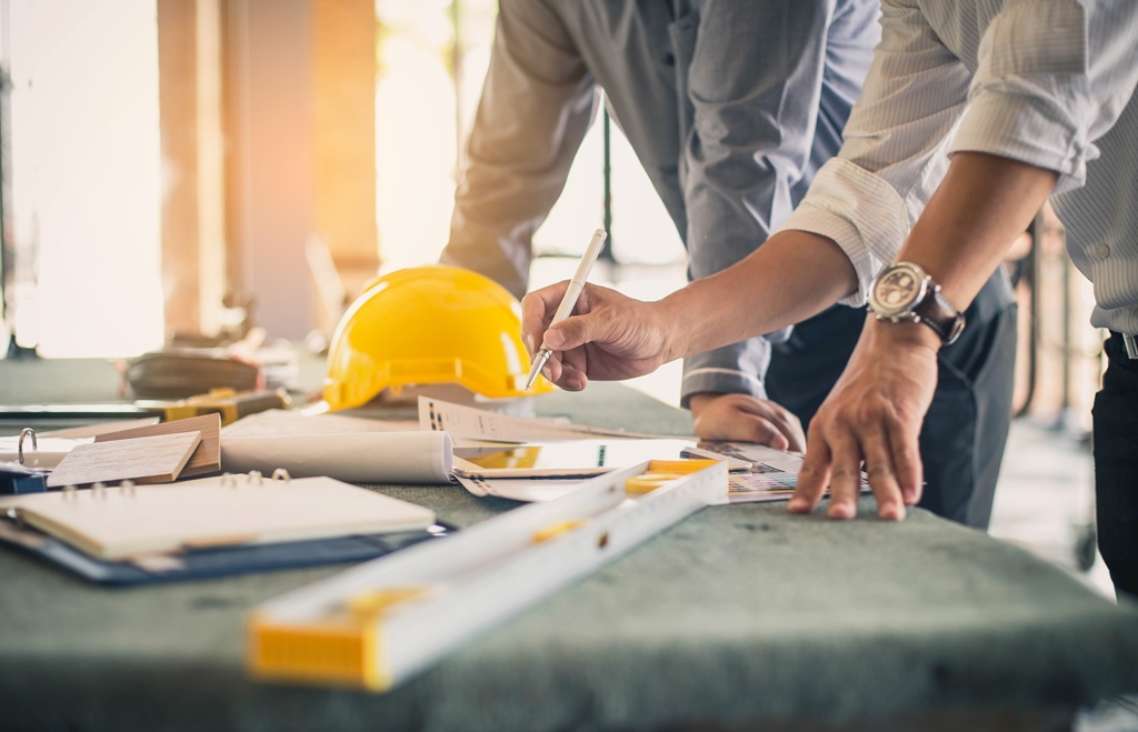 Two professionals working on architectural plans with a hard hat and tools on the table.